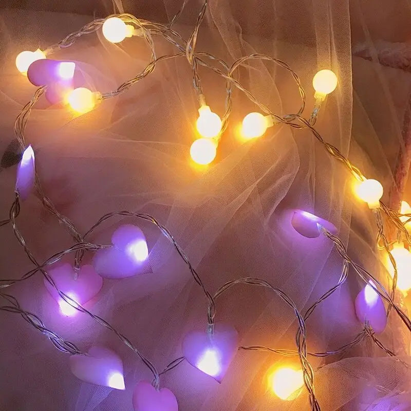 String lights with glowing yellow bulbs and purple heart-shaped covers arranged on soft pink fabric, creating a warm and cozy ambiance.