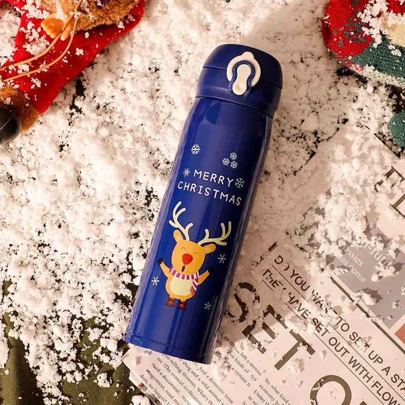 Blue thermos bottle with a festive reindeer and "Merry Christmas" design on a snowy surface. Scattered holiday decorations and a partially visible newspaper add a seasonal touch.