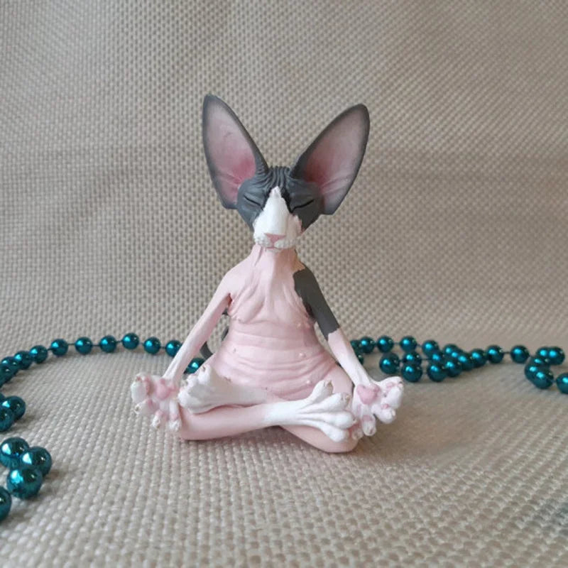A small figurine of a hairless cat sitting in a yoga pose, with eyes closed and paws in a meditative position. It is surrounded by a strand of turquoise beads on a beige fabric.