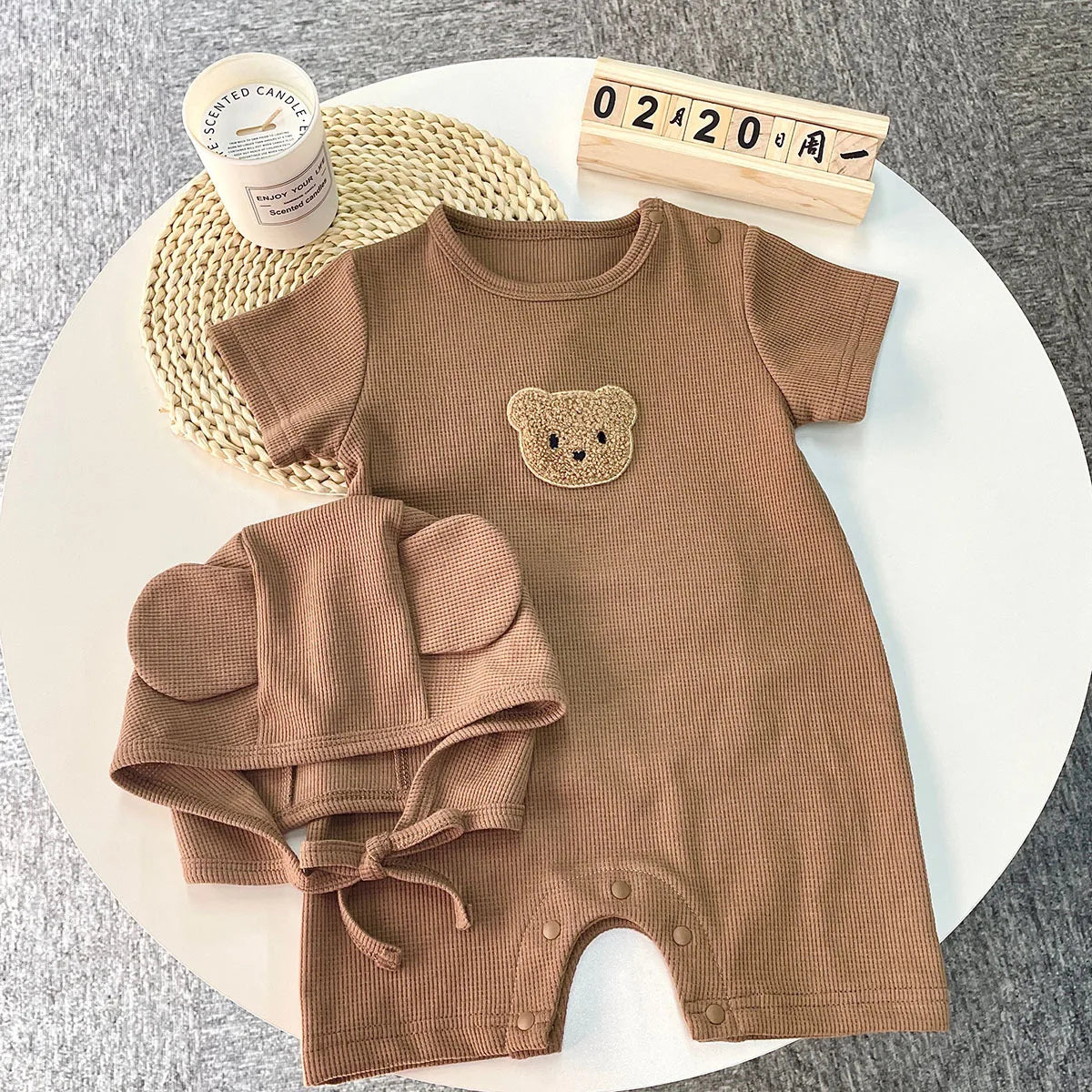 Brown baby romper with a bear patch and matching hat with ears, laid on a white table with a calendar block showing 0220 and a scented candle nearby.