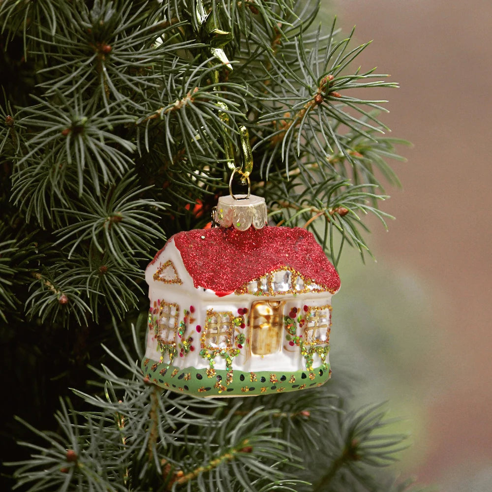 Christmas tree ornament shaped like a house with a glittery red roof and colorful decorations, hanging on a lush green pine branch.