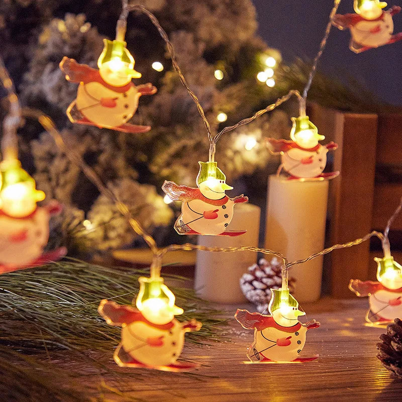 Festive string lights feature glowing snowmen with red scarves and hats, illuminating a cozy setting with candles, pine branches, and a softly lit tree in the background.