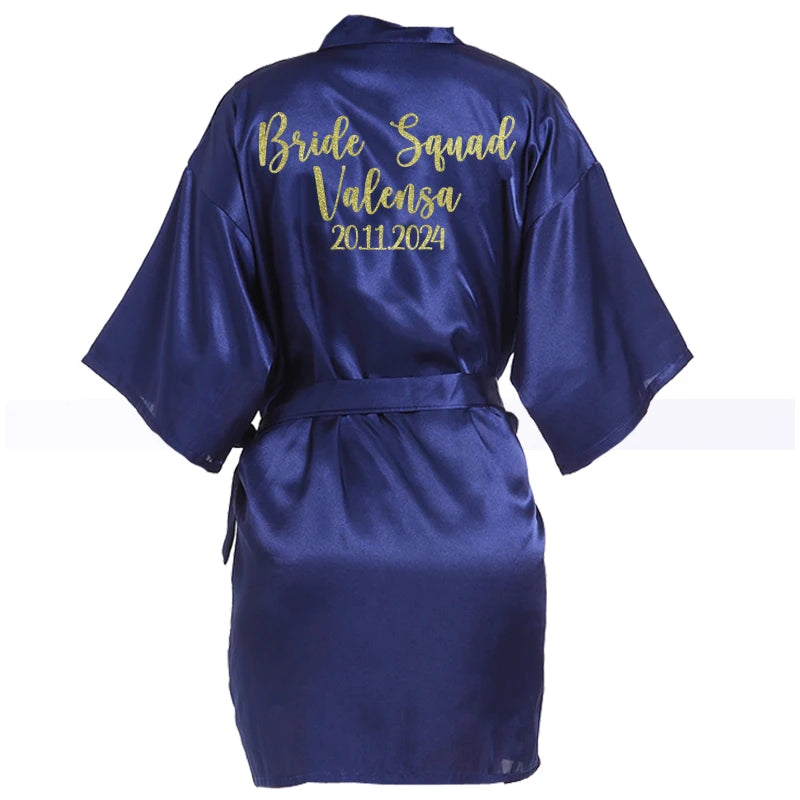 Navy blue satin robe with "Bride Squad Valensa 20.11.2024" embroidered in gold on the back. Features short sleeves and a tie belt at the waist.