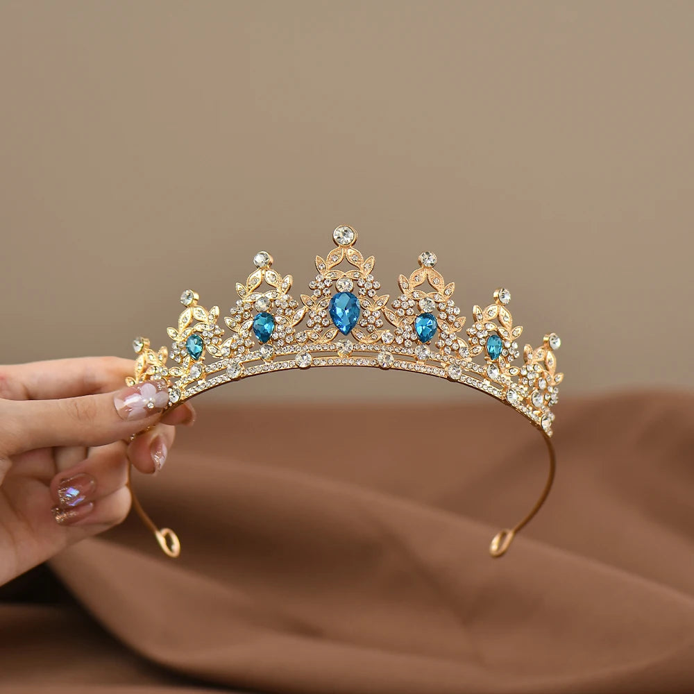 A person holds an ornate golden tiara adorned with blue gemstones and small diamonds, set against a soft brown fabric background.