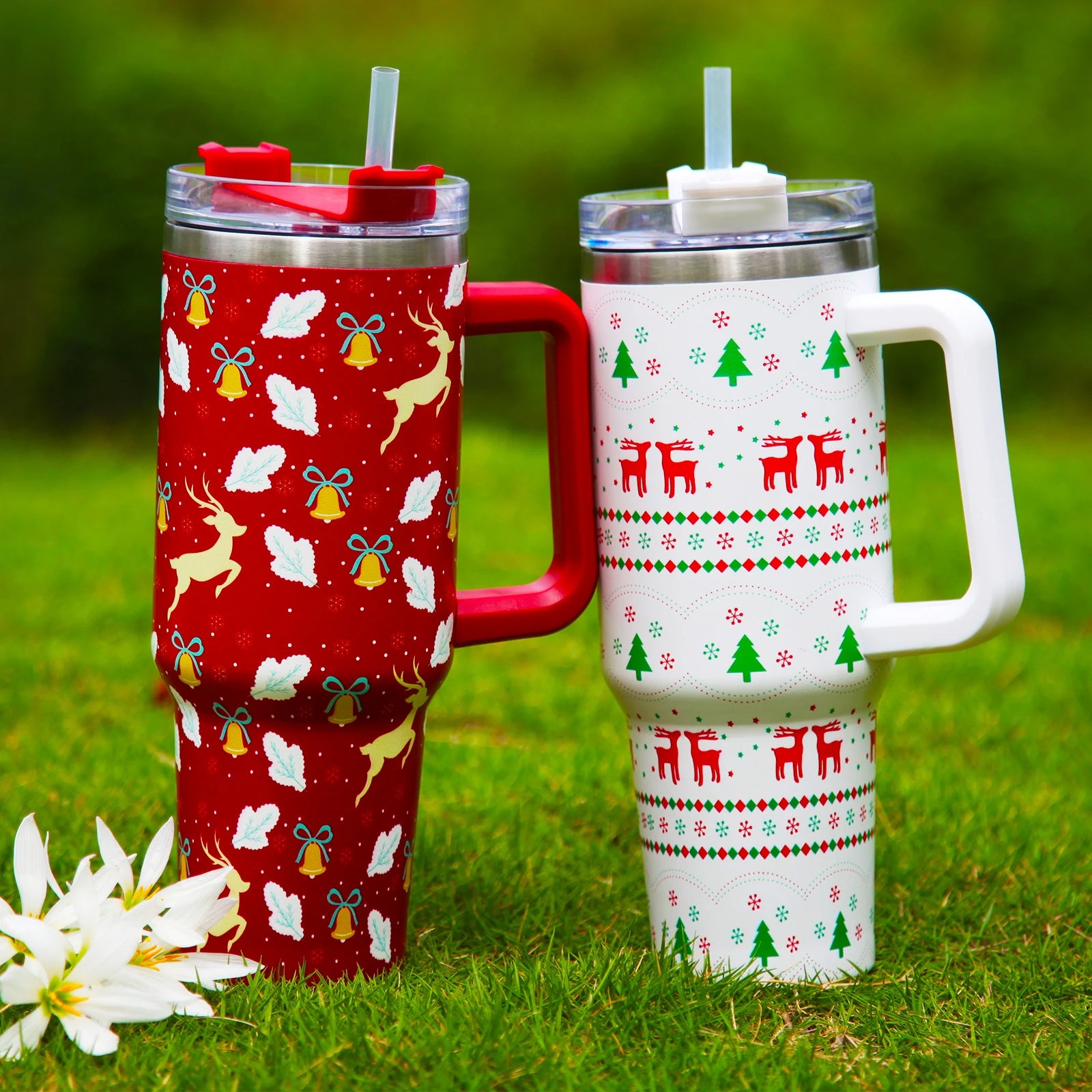 Two festive travel mugs on grass; one red with reindeer, bells, leaves, and snowflakes, and another white with red reindeer, green trees, and geometric patterns, both with lids and straws.