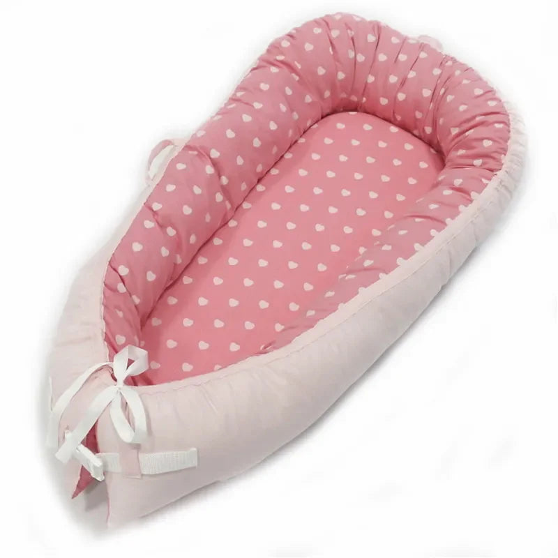 Pink baby nest with a pattern of white hearts, featuring soft padded edges and tied with white ribbons at the bottom, providing a cozy sleeping space for infants.