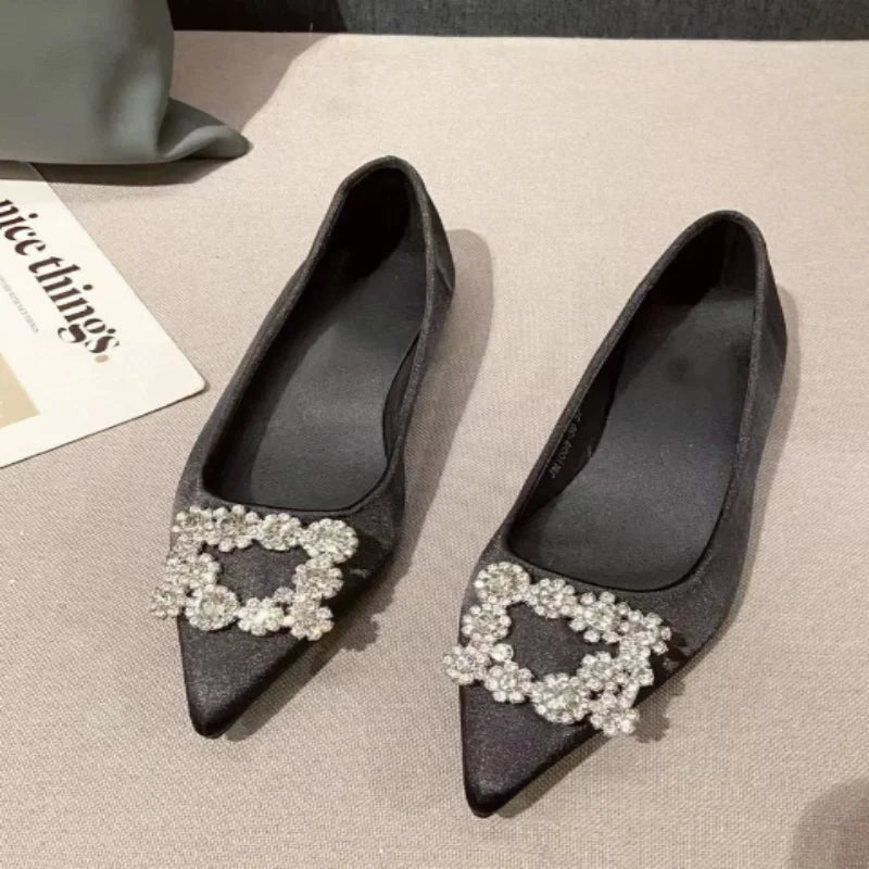 Black pointed-toe flats with sparkling floral embellishments on the front. They are displayed on a beige surface with part of a magazine visible.