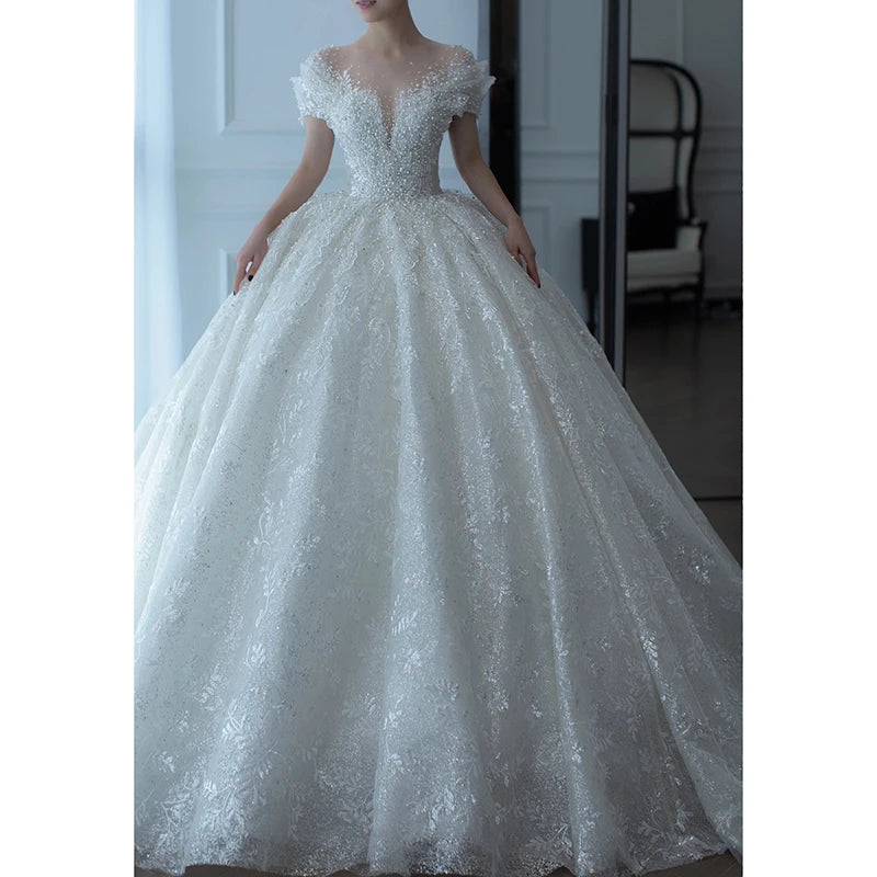 A bride in a voluminous white lace ball gown with a deep V-neckline and cap sleeves, standing in a softly lit room with white walls and elegant furniture.