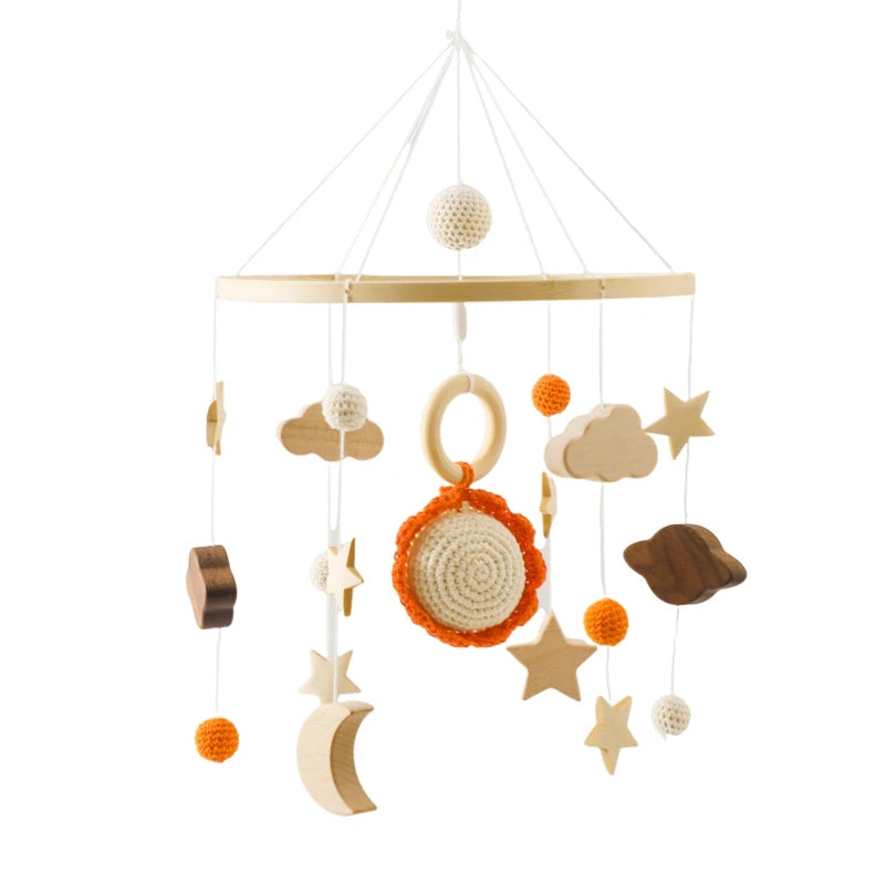A baby mobile with wooden and crocheted elements, featuring stars, clouds, a sun, a moon, and decorative balls in white, orange, and brown, hanging from a circular frame.