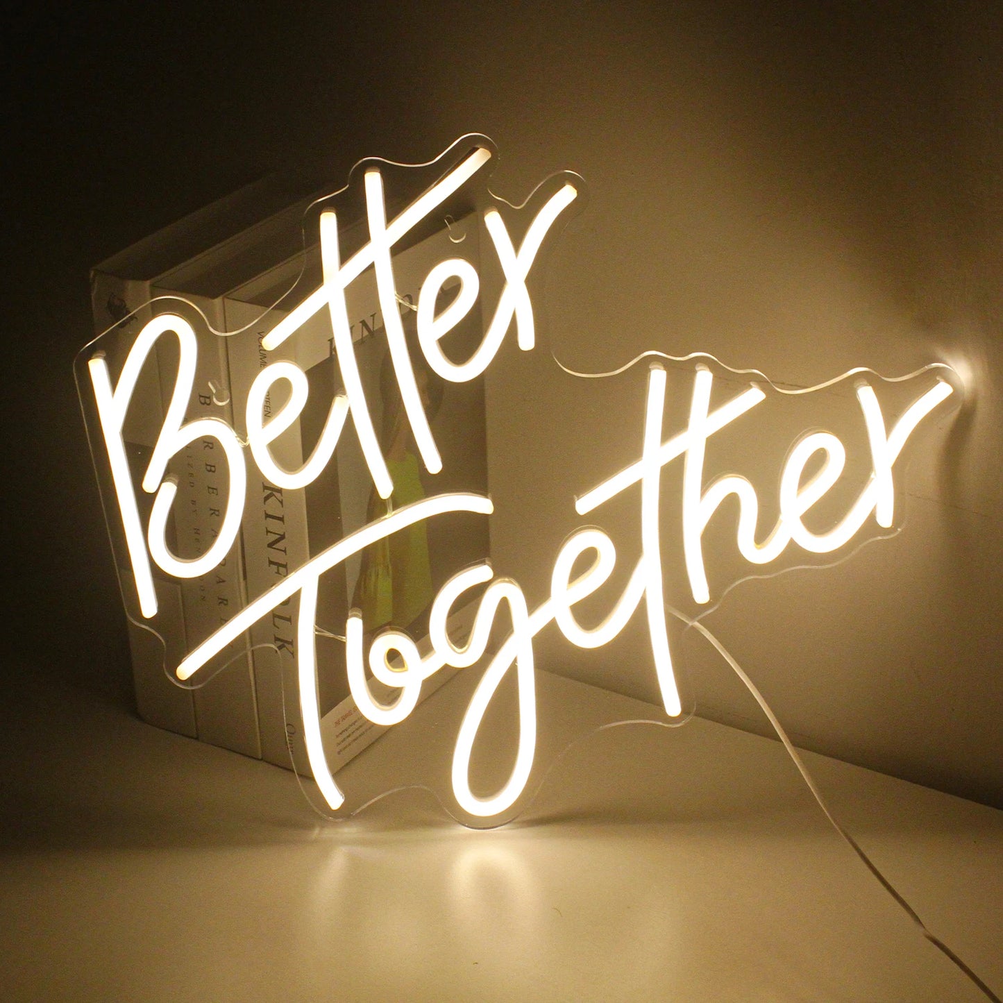 Illuminated neon sign reading "Better Together" in a cursive script, placed against a wall. The warm glow creates a cozy ambiance, partially illuminating books behind it.