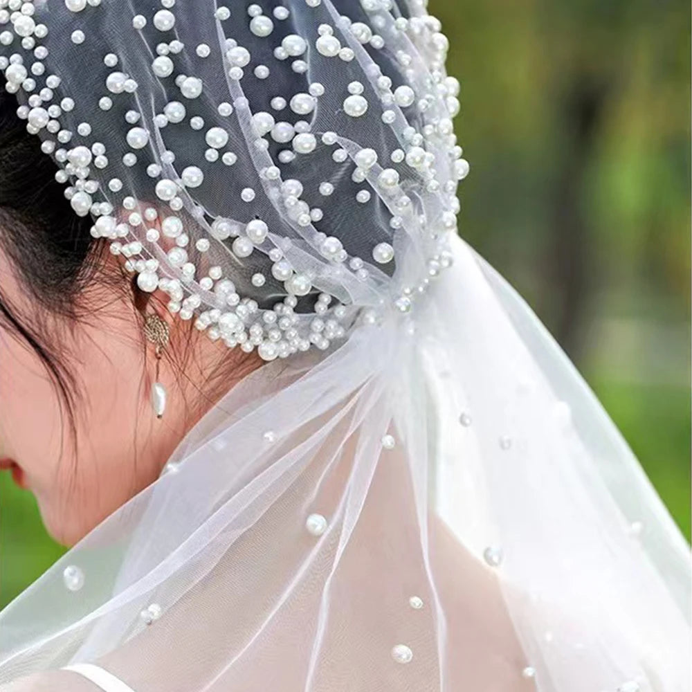 Close-up of a person's head wearing a delicate veil adorned with white pearls. The veil drapes gracefully over dark hair, creating an elegant and sophisticated look.