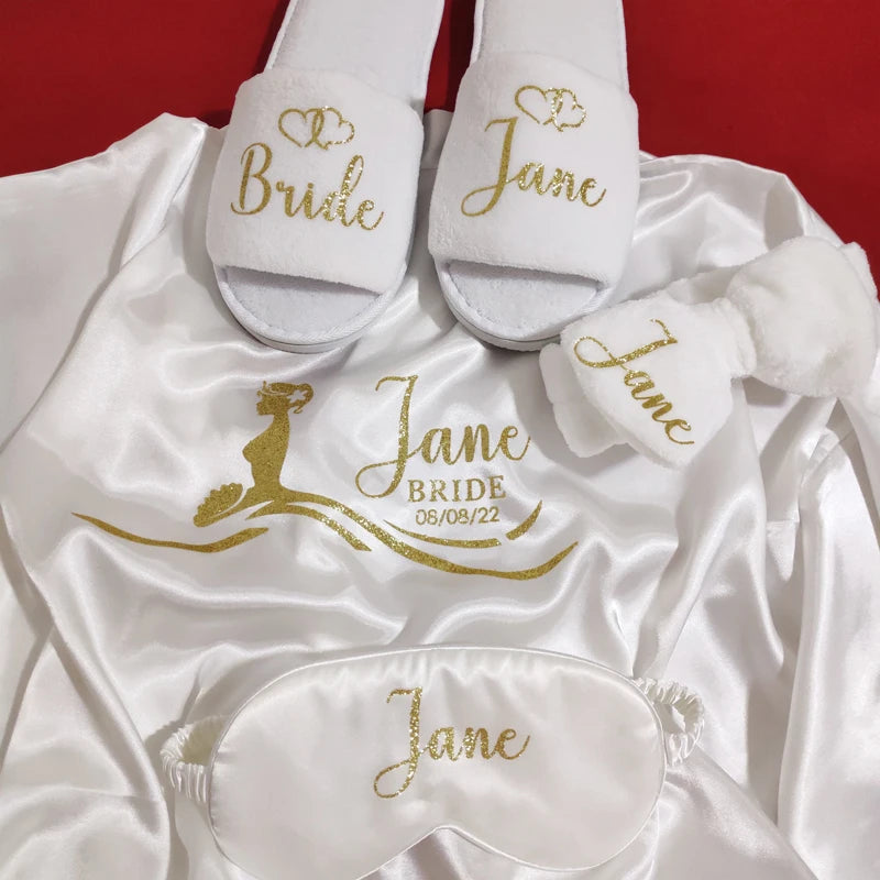 Bridal-themed items on a satin fabric: slippers and a robe with "Bride" and "Jane," a sleep mask, and headband, all in gold text. Date 08/08/22 is on the robe.