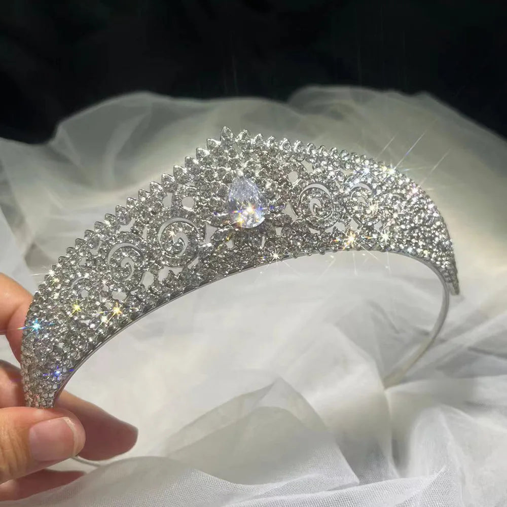 A sparkling silver tiara adorned with intricate diamond patterns and a central teardrop gem is held against a soft white fabric background, catching glimmers of light.
