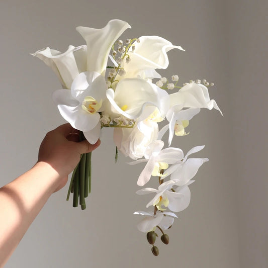 A hand holds a bouquet of elegant white flowers, including calla lilies and orchids, against a neutral background, with soft light illuminating the petals.