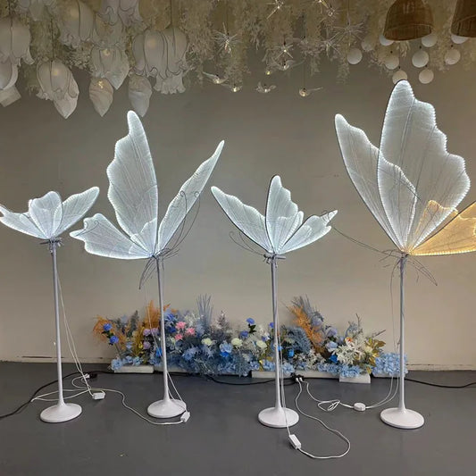 Four floor lamps with illuminated butterfly wings in varying heights stand in front of a floral backdrop. The lamps cast a soft glow, enhancing the whimsical decor.