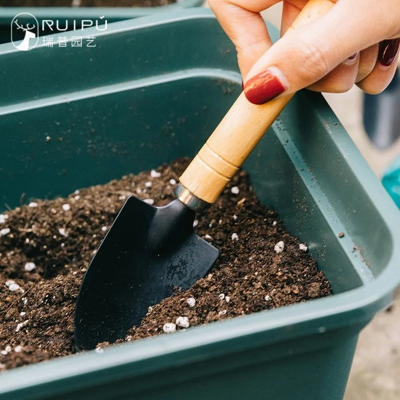 Close-up of a hand with red nails holding a small gardening trowel, digging into dark soil in a green container. The trowel has a wooden handle and a black metal blade.