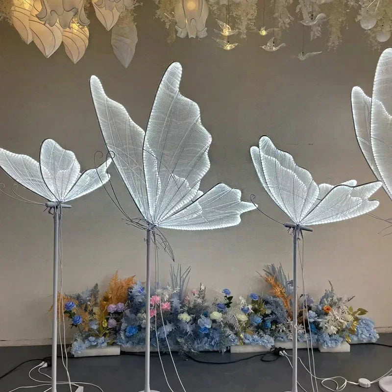 Illuminated butterfly sculptures with translucent wings glow against a neutral backdrop. Beneath, a display of soft blue and pastel flowers adds a touch of color.