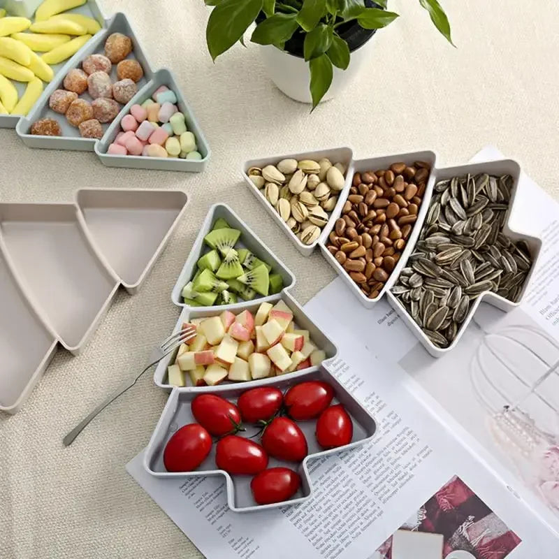 Tree-shaped plates filled with sliced fruit, nuts, seeds, tomatoes, and sweets are arranged on a table. A plant and open book are placed nearby.