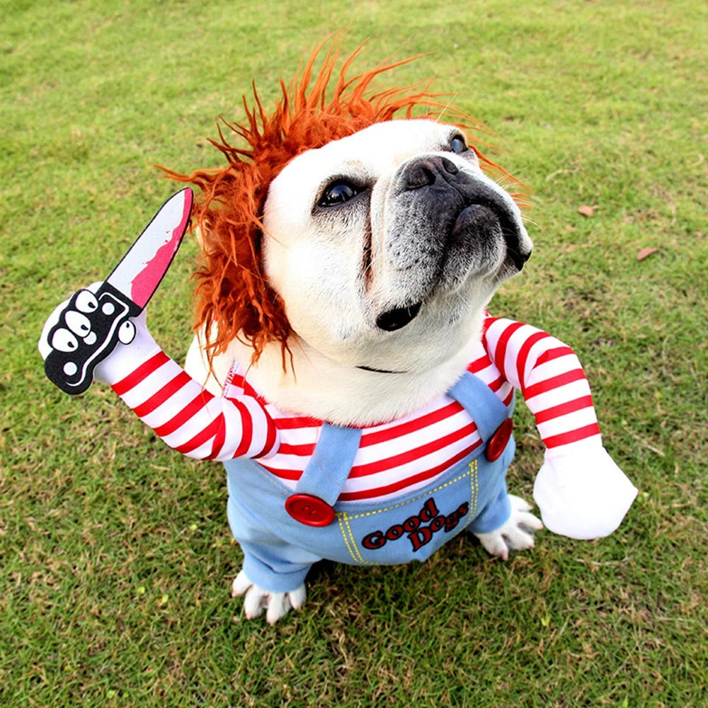Bulldog in a playful costume stands on grass, wearing a red wig and striped outfit with faux arms holding a toy knife. The costume says "Good Dog" on blue overalls.