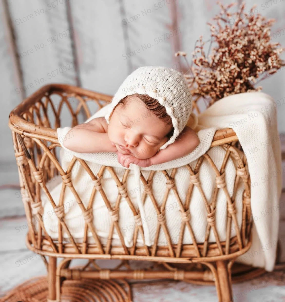 Newborn baby sleeping peacefully in a wicker basket, wearing a knitted cap. The baby is swaddled in a white blanket, with dried flowers in the background.