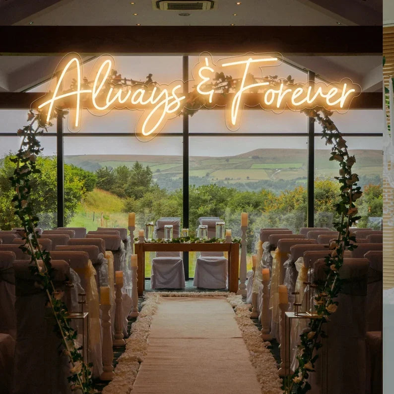 Wedding ceremony venue with chairs arranged in rows, a floral arch, and a neon sign reading "Always & Forever" against glass windows showcasing a scenic countryside view.