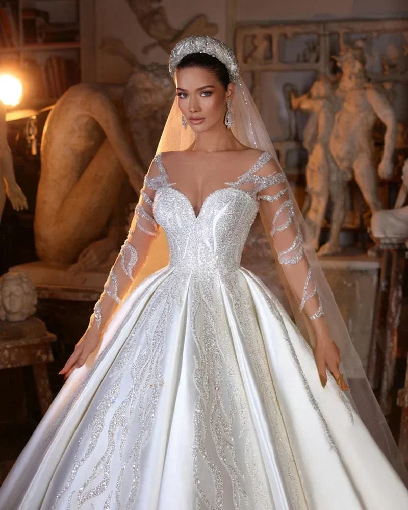 Bride in an opulent white gown with intricate beading and sheer long sleeves, wearing a sparkling headpiece and veil. Sculptures and soft lighting create an artistic backdrop.
