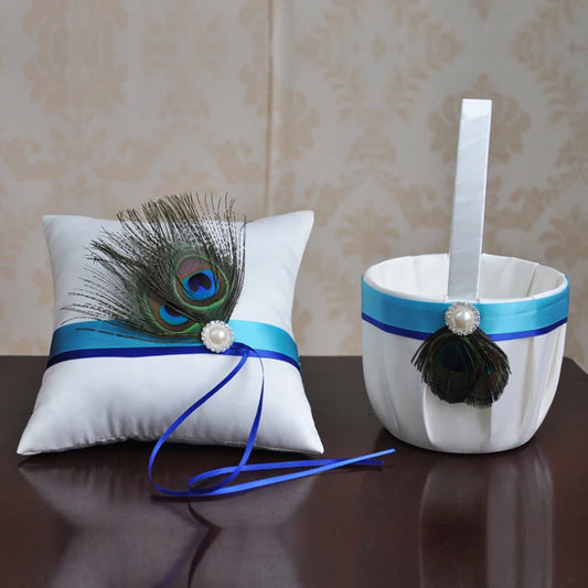 White satin ring bearer pillow and basket, both adorned with blue ribbon, pearls, and peacock feathers, placed on a dark wooden table against a patterned wall.
