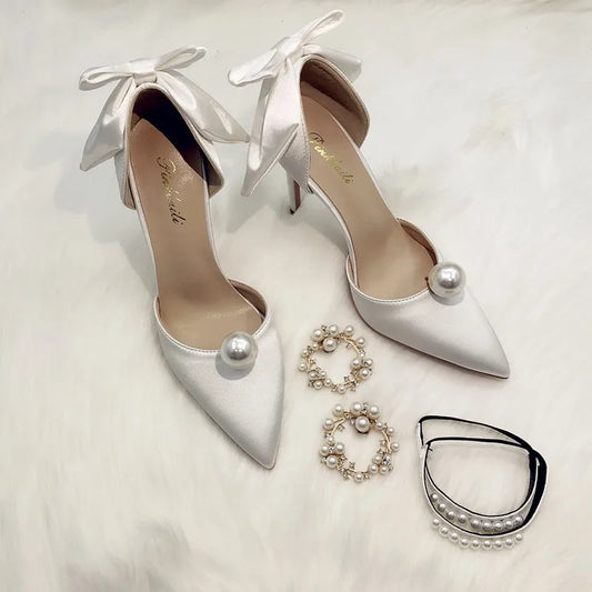 Elegant white stiletto heels with pearl accents and satin bows, paired with pearl hoop earrings and a pearl-accented choker on a soft white surface.