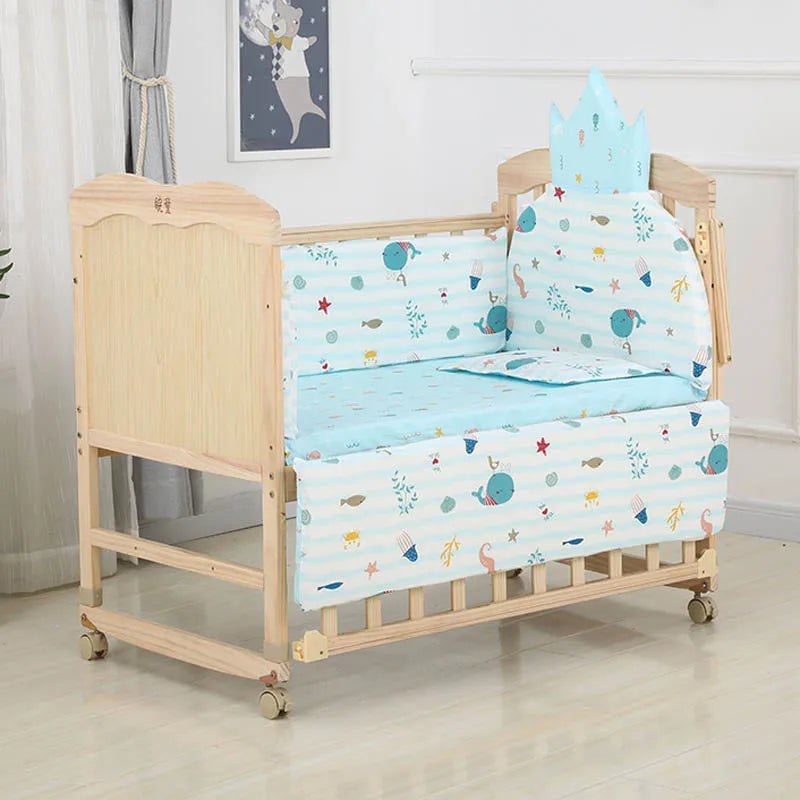 Wooden baby crib with blue ocean-themed bedding, including whales and fish. Features a crown-shaped headboard cushion. Room has light walls, a curtain, and a moon-themed wall picture.