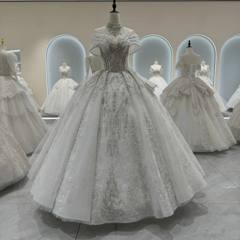 Elegant white ball gown adorned with intricate silver embroidery and sparkling details, displayed on a mannequin. Other similar gowns are visible in the softly-lit showroom.