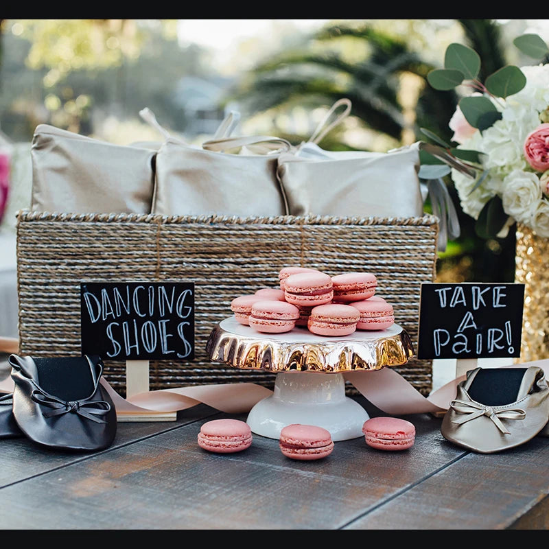 A wicker basket with satin bags, pink macarons on a cake stand, and ballet flats titled "Dancing Shoes." Signs read "Take a Pair!" amid lush greenery and flowers.