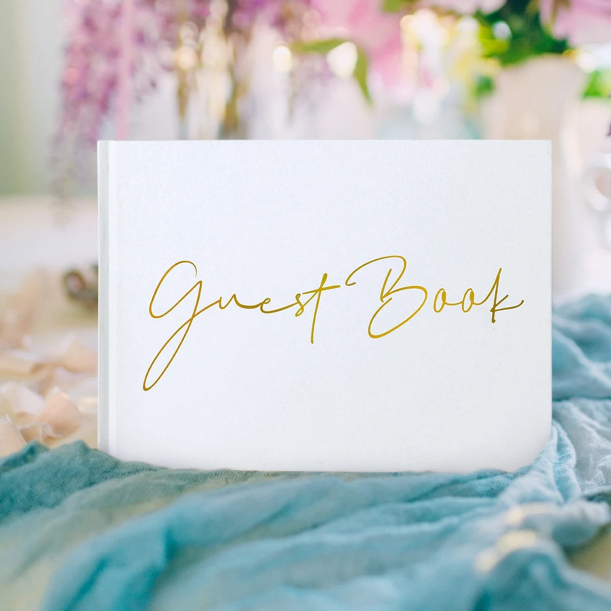 Elegant guest book with gold script on a white cover, placed on a table with soft blue fabric and pastel flowers in the background.