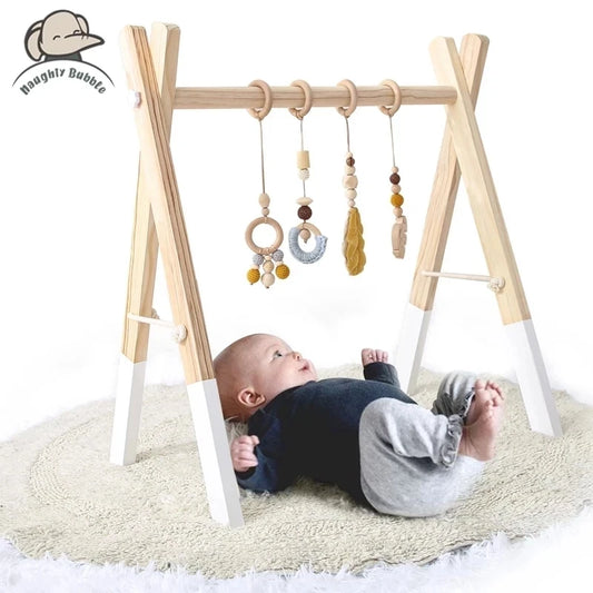 Baby lying on a cream rug, gazing up at a wooden baby gym with hanging toys. The gym includes rings, beads, and textured shapes in natural and yellow hues.
