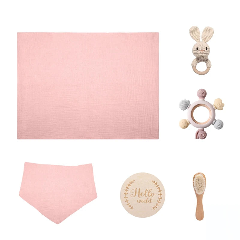 Pink baby gift set with a soft blanket, bib, crochet bunny rattle, wooden "Hello World" plaque, pastel teething ring, and natural bristle brush on a white background.