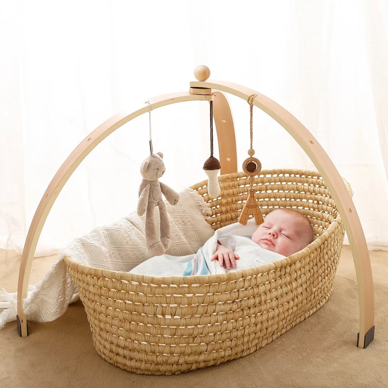 A sleeping baby lies in a woven bassinet under a wooden arch adorned with hanging toys, including a teddy bear and Eiffel Tower. The background is softly lit with curtains.