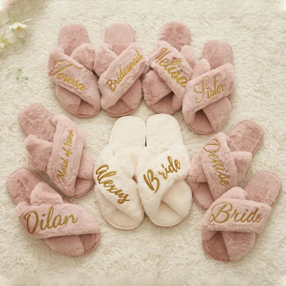 A collection of fluffy slippers in pink and white arranged in a circle on a soft carpet. Each pair has a name or title embroidered in gold, including "Bride" and "Bridesmaid."