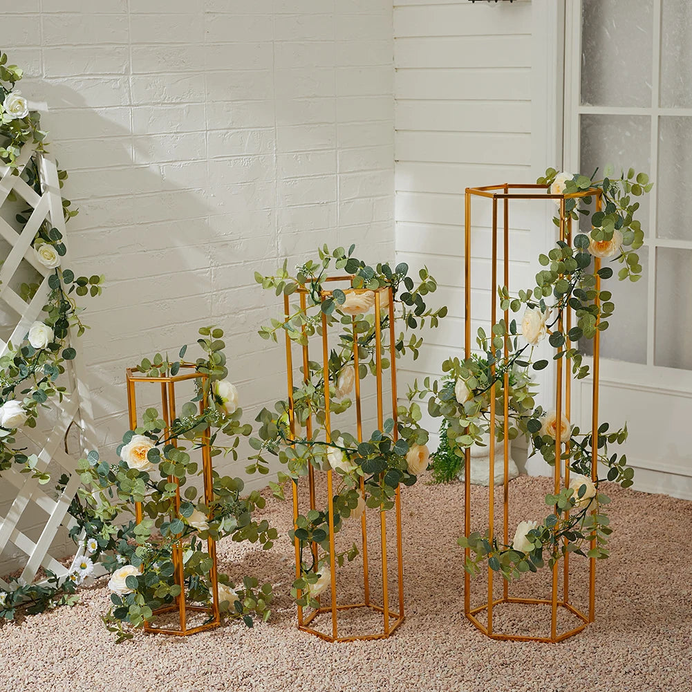 Three gold geometric plant stands with green and white floral arrangements against a white brick wall and window, set on a pebble-textured floor.