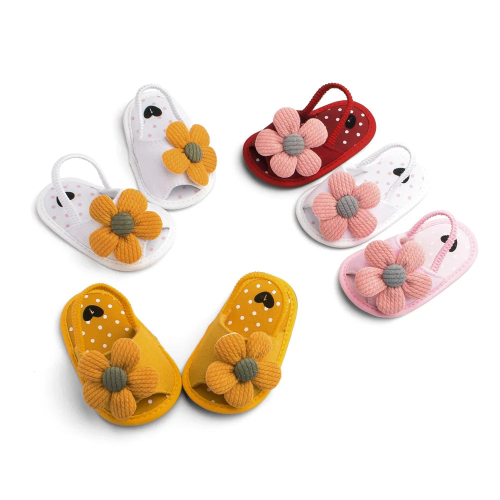 Colorful baby sandals with large fabric flowers. Designs include white with yellow flower, red with pink flower, yellow with yellow flower, and pink with pink flower on a white background.