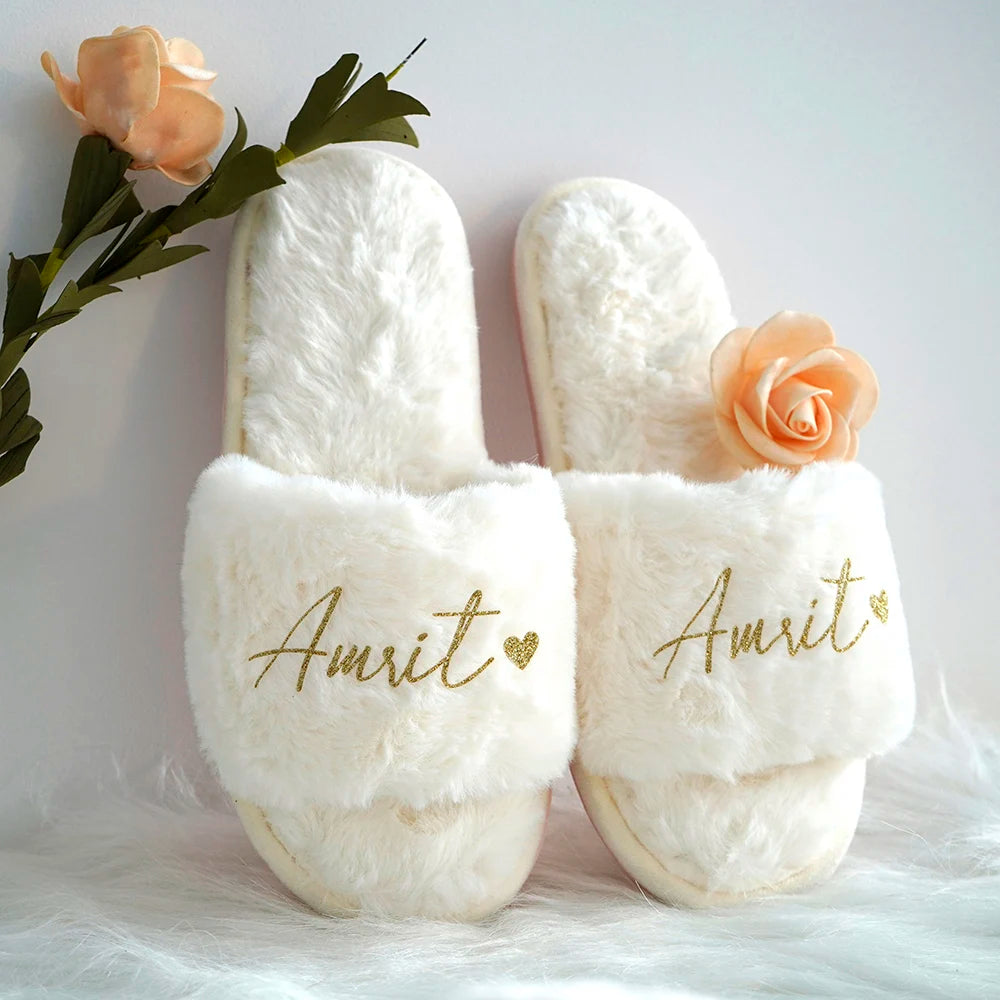 Fuzzy white slippers with gold script on the straps. One slipper has a peach rose tucked in, and they're placed near a similar rose on a fluffy white surface.