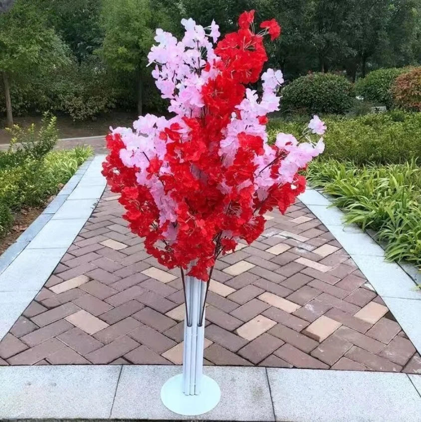Artificial red and pink flower arrangement in a metallic vase placed on a herringbone brick path, surrounded by lush greenery in a garden setting.