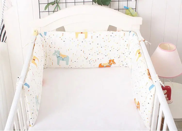 White crib with colorful animal-patterned bumpers and a white mattress. A pink lamp and green plant in the background create a soft, cozy nursery setting.