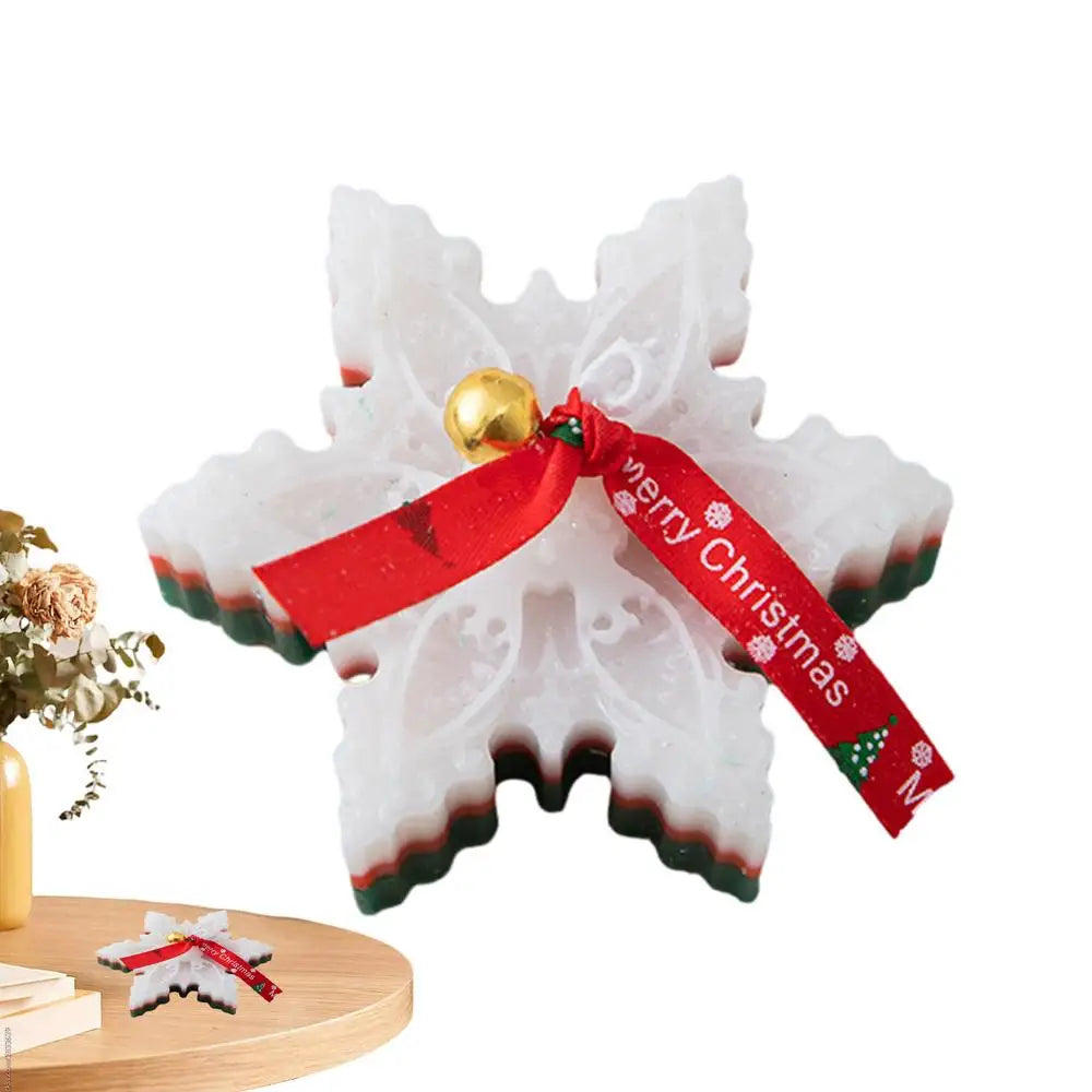A snowflake-shaped soap ornament with white, red, and green layers. It features a red ribbon with "Merry Christmas" and a gold bell, placed on a wooden surface near flowers.