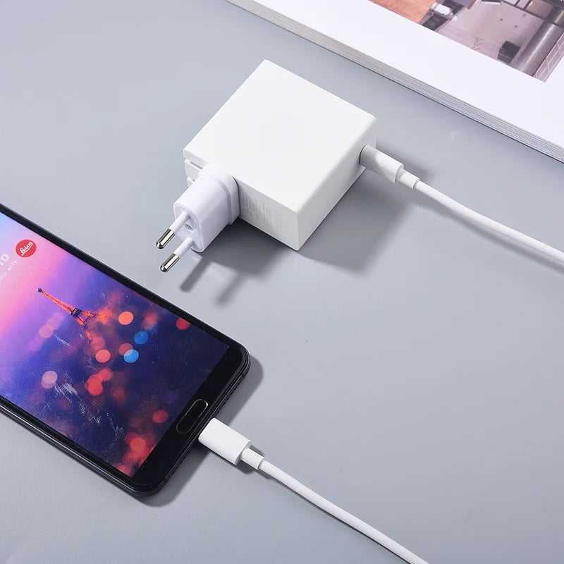 A smartphone is charging with a white plug adapter and USB cable on a gray surface, displaying a colorful wallpaper with a tower silhouette. A book lies nearby.