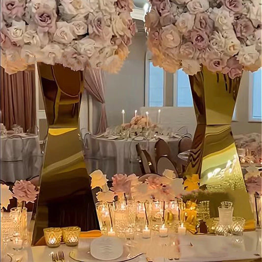 Elegant dining table setting with large golden vases topped with pink and white roses. Candles and glasses create a warm ambiance. Luxurious drapes and chairs in the background.