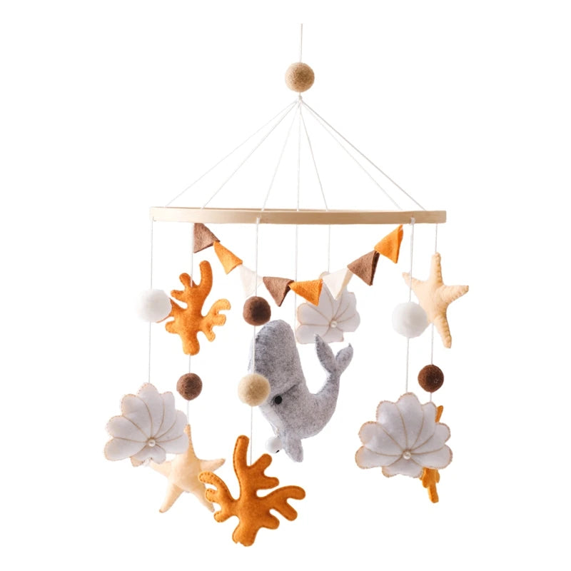 A baby mobile featuring felt sea creatures including a gray whale, orange coral, tan starfish, and white seashells, hanging from a hoop with small flags and pom-poms.