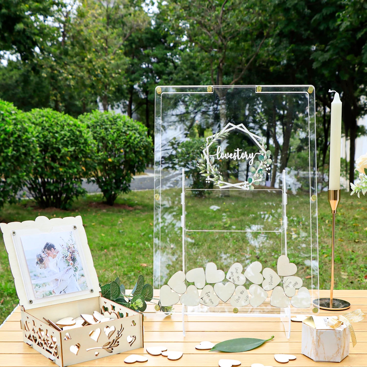 Clear wedding card box with heart-shaped notes on a wooden table, surrounded by greenery. There's a photo frame and a tall candle nearby, creating a romantic outdoor setting.