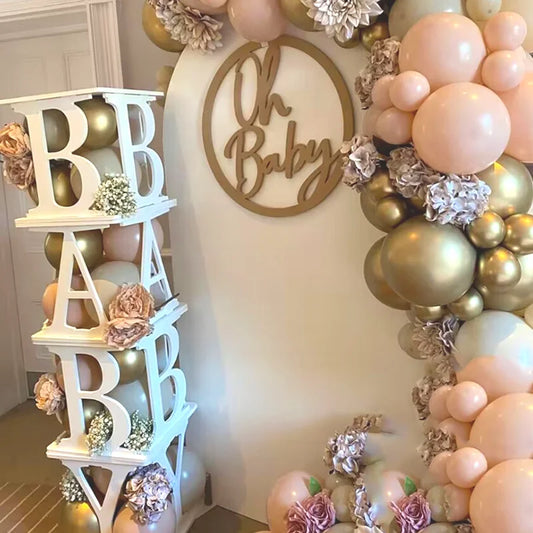 Baby shower decor with gold, cream, and pink balloons forming an arch. A sign reads "Oh Baby" and a block tower spells "BABY" with floral accents.