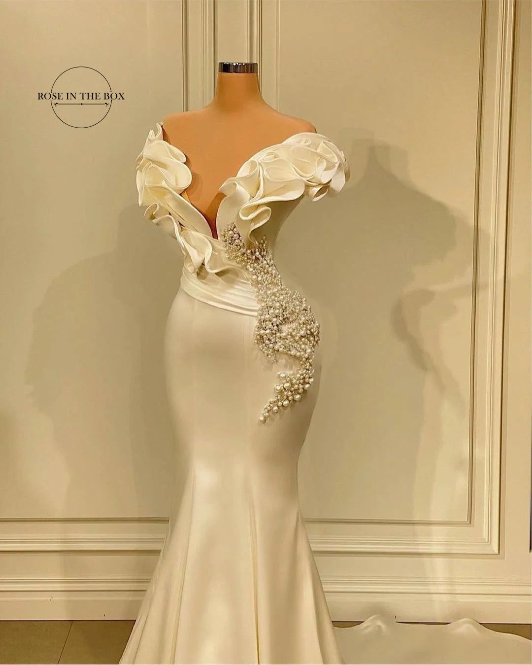 Elegantly designed off-shoulder ivory gown on a mannequin with ruffled floral detailing and pearl embellishments cascading down the side.