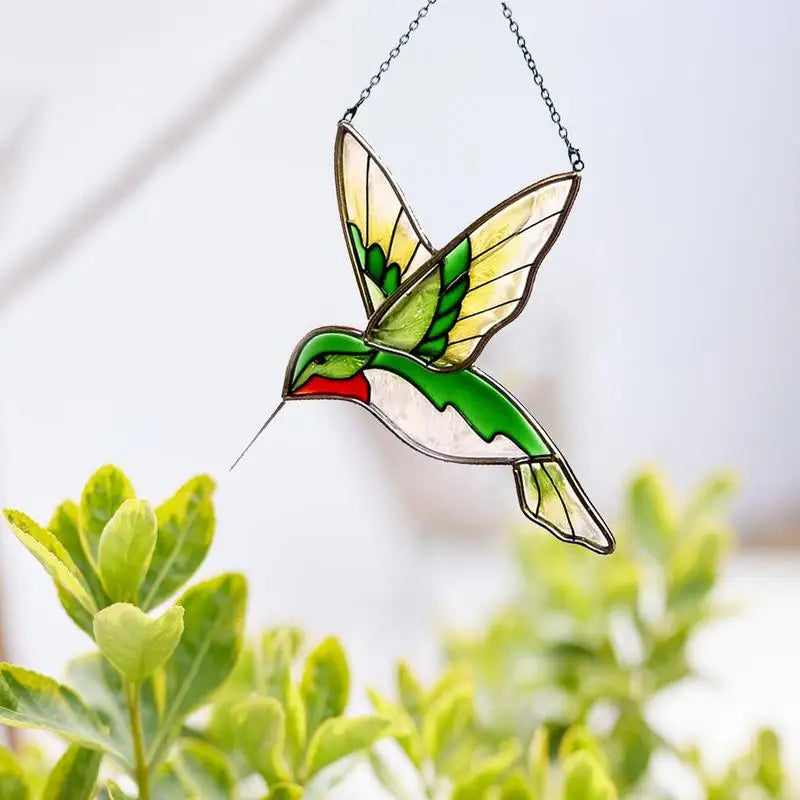 Stained glass hummingbird decoration with green and yellow wings, hanging in front of leafy plants, against a light background.