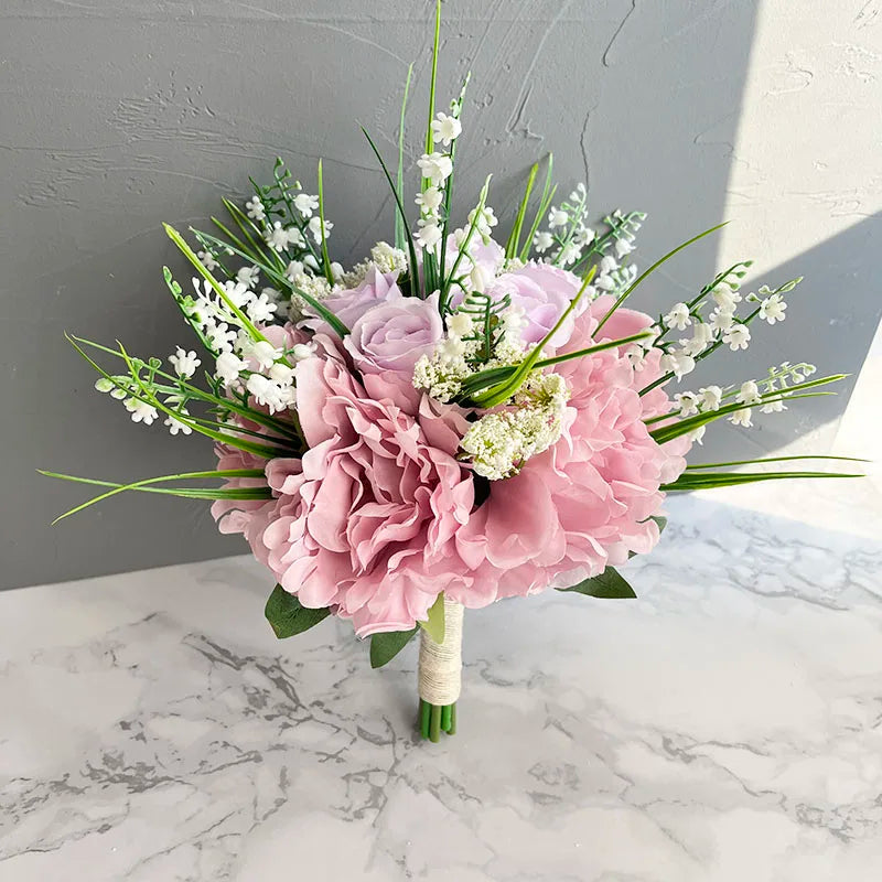 A bouquet of pink hydrangeas, lavender roses, and white lilies with green leaves, wrapped in white ribbon, sits on a marble surface against a gray background.